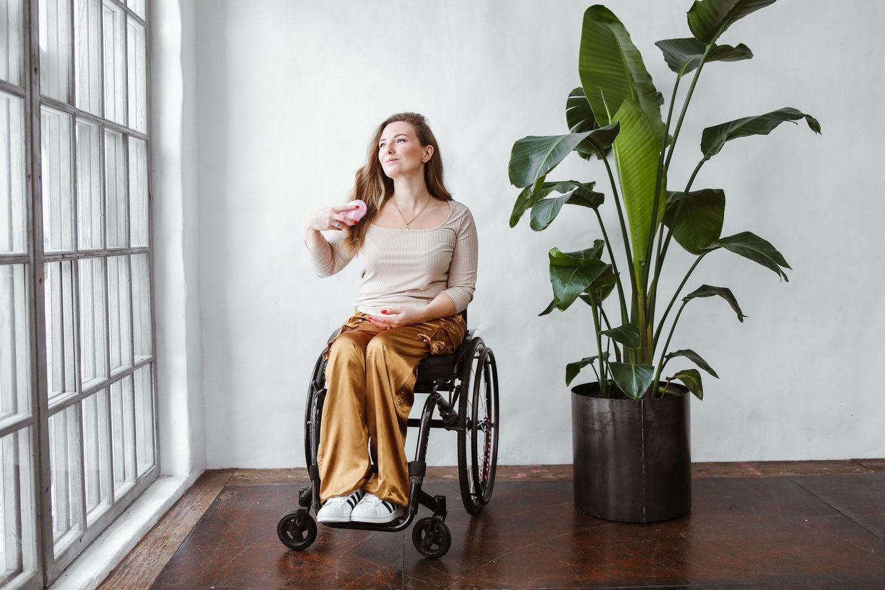 A woman using a wheelchair brushes her hair beside a window.